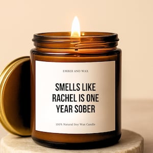 May include: A burning brown glass jar candle with a gold lid, on a stone coaster. The label reads "SMELLS LIKE RACHEL IS ONE YEAR SOBER." The text "EMBER AND WAX" and "100% Natural Soy Wax Candle" are also visible.