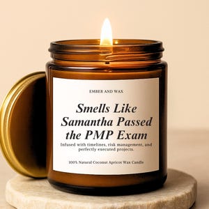 Puede incluir: Una vela encendida en un frasco de vidrio marrón con tapa dorada. La etiqueta blanca dice: Ember and Wax, Smells Like Samantha Passed the PMP Exam, Infused with timelines, risk management, and perfectly executed projects, 100% Natural Coconut Apricot Wax Candle.