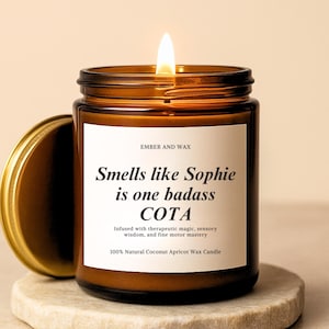 May include: A lit brown glass candle jar with a gold lid, sitting on a stone coaster. The label reads "Smells like Sophie is one badass COTA" and "100% Natural Coconut Apricot Wax Candle."