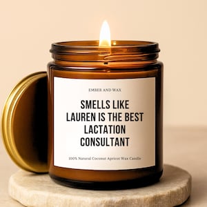 May include: A lit brown glass candle jar with a gold lid, sitting on a stone coaster. The candle has a white label with the text "SMELLS LIKE LAUREN IS THE BEST LACTATION CONSULTANT". The candle is made of 100% Natural Coconut Apricot Wax.