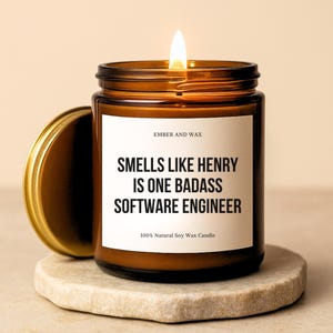 Peut inclure: Une bougie en cire de soja allumée dans un pot en verre marron avec un couvercle doré. L'étiquette indique "SMELLS LIKE HENRY IS ONE BADASS SOFTWARE ENGINEER". La bougie est posée sur un dessous de verre en pierre. Le texte "EMBER AND WAX" est également visible.