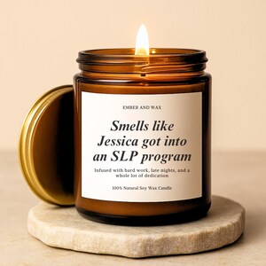 May include: A lit, brown glass candle jar with a gold lid, sitting on a stone coaster. The candle has a white label with the text "Smells like Jessica got into an SLP program." The candle is made of 100% natural soy wax.