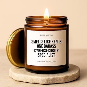 May include: A lit, brown glass candle jar with a gold lid, on a stone coaster. The candle has a white label with the text "SMELLS LIKE KEN IS ONE BADASS CYBERSECURITY SPECIALIST" and "100% Natural Soy Wax Candle".
