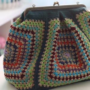 May include: A handmade crochet purse with a metal clasp. The bag features a colorful granny square design in shades of blue, green, orange, purple, and gold. The purse has a dark teal border and a vintage-style clasp.