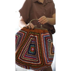 Op de afbeelding: Een kleurrijke, handgemaakte gehaakte tas met een bruin frame en handvat. De tas heeft een geometrisch patroon in verschillende kleuren, waaronder oranje, blauw, groen en wit. De tas wordt vastgehouden door een persoon die een bruine outfit draagt.