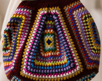 May include: A colorful, handmade crochet bag with a dark brown frame and clasp. The bag features a vibrant patchwork design with squares of various colors, including blue, yellow, green, and purple. The bag has a metal clasp and a chain strap.