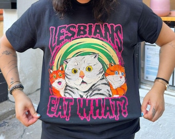 May include: A black t-shirt with a graphic print of a cat with the text "Lesbians Eat What?" in pink. The cat is surrounded by a green and white swirl. The shirt also has the text "high-end gay." in yellow on a black baseball cap.