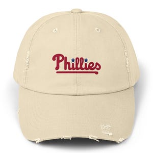 Könnte beinhalten: Eine beige, abgenutzte Baseballkappe mit dem Wort "Phillies" in rot gestickt, darüber zwei blaue Sterne.