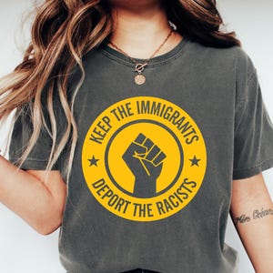 May include: Dark gray t-shirt with a yellow circular graphic. The graphic features a raised fist inside a circle with the text "KEEP THE IMMIGRANTS" above and "DEPORT THE RACISTS" below. Two stars flank the fist.