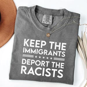 May include: A grey Comfort Colors t-shirt with the text "KEEP THE IMMIGRANTS DEPORT THE RACISTS" in white. A gold necklace and a brown hat are also visible. The shirt is folded and lies on a white surface.