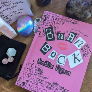 May include: A pink paperback book titled "Burn Book" by Nadia Lynn. The cover features black and white letters with a red lipstick kiss mark. The cover is decorated with black and white doodles.