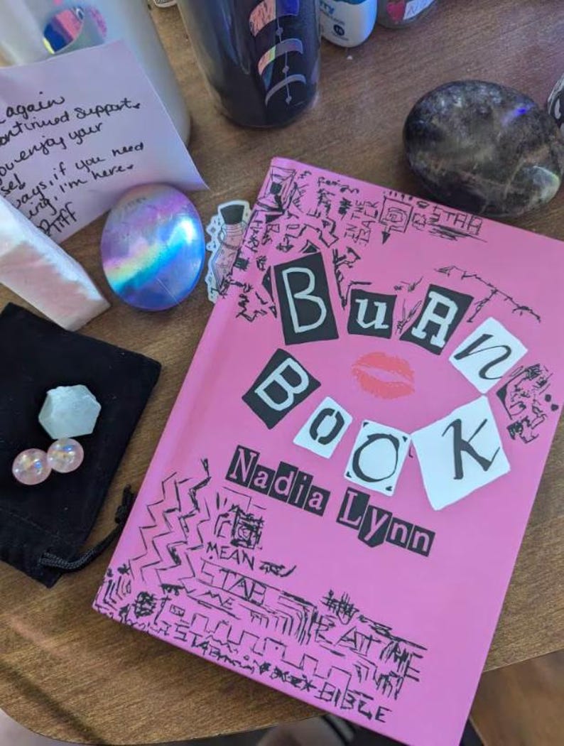 May include: A pink paperback book titled "Burn Book" by Nadia Lynn. The cover features black and white letters with a red lipstick kiss mark. The cover is decorated with black and white doodles.