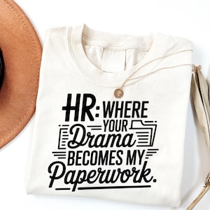 Könnte beinhalten: Cremefarbenes T-Shirt mit dem schwarzen Text "HR: Where Your Drama Becomes My Paperwork". Eine goldene Halskette mit Anhänger liegt auf dem Shirt. Ein brauner Hut ist in der oberen linken Ecke zu sehen.