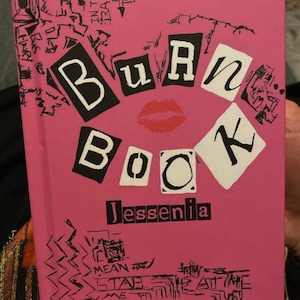 May include: Pink book with the title "Burn Book" in black letters with a red lip print in the center. The book is written by "Jessenia" in black letters.