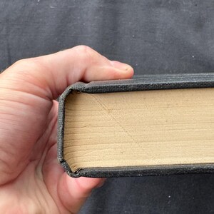 May include: A hardcover book with a dark gray textured fabric cover and light tan pages. The spine edge of the book shows a distinct diagonal crease mark across the page edges, indicating the book has been opened or bent at that specific point.