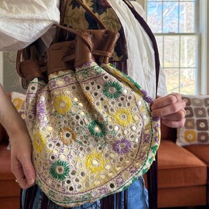 May include: A cream-colored drawstring bag with embroidered floral patterns in yellow, green, and purple, accented with small mirrors. The bag has brown leather straps and a fringe detail.