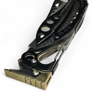 May include: A black multi-tool with a gold-colored handle and a carabiner clip. The tool has a variety of blades and tools, including a knife, a saw, a screwdriver, and a pliers. The tool is made by Leatherman and is called the "Sidekick CX".