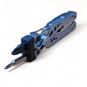 Puede incluir: Una multiherramienta plateada con un portapuntas negro y dos puntas. La herramienta tiene alicates, un cuchillo y otros implementos. La herramienta está diseñada para diversas tareas y es compacta para facilitar su transporte.