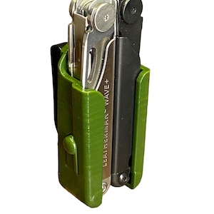 Könnte beinhalten: Ein grünes Kunststoffholster mit einem schwarzen und silbernen Multitool im Inneren. Das Multitool ist mit "LEATHERMAN WAVE+" beschriftet.