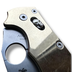 Puede incluir: Primer plano de una navaja de bolsillo dorada con un mango en forma de calavera. La navaja tiene una hoja negra y un logotipo de Spyderco en el lateral.