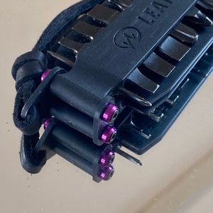 May include: A black multi-tool with a leather strap and purple screws. The tool has a variety of blades and tools.