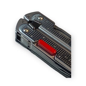 Könnte beinhalten: Ein silbernes Multitool mit einem roten Knopf. Das Werkzeug hat eine strukturierte Oberfläche und eine Vielzahl von Werkzeugen.