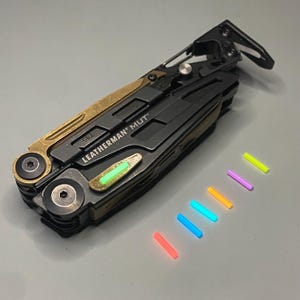 Könnte beinhalten: Ein schwarzes und goldenes Multitool mit dem Text "LEATHERMAN MUT" an der Seite. Das Werkzeug ist geöffnet und hat ein grünes Licht an der Seite. Auf einer grauen Oberfläche liegen sieben Leuchtstäbe in verschiedenen Farben.