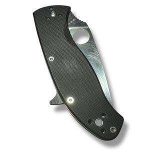 Peut inclure: Un couteau pliant noir avec une lame partiellement ouverte. Le manche a une surface texturée et plusieurs vis argentées.