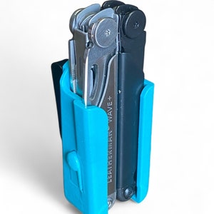 Könnte beinhalten: Ein Multitool mit schwarzem Griff und silbernen Werkzeugen wird in einer leuchtend blauen Hülle gehalten. Der Text "LEATHERMAN WAVE+" ist auf dem Werkzeug sichtbar. Die Hülle hat einen Clip.