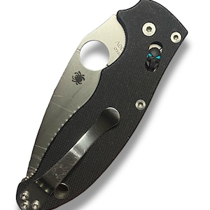 Peut inclure: Un couteau pliant noir avec une lame argentée et un clip de poche en forme de crâne. La lame porte le logo Spyderco et le texte "154CM".