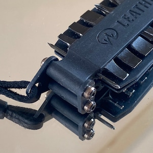 Peut inclure: Un multi-outil Leatherman noir avec une lame argentée et un cordon noir. L'outil possède divers accessoires, dont un tournevis, un couteau et une lime.