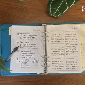 May include: A blue and white planner with a monthly check-in section, notes section, and a list of "quick wins" and "high priority" tasks. The planner has a tabbed section with the numbers 1 through 6. The text "Plannable Human" is printed in the upper right corner of the page.