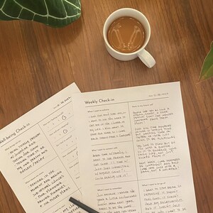 May include: Two printable worksheets on a wooden table with a cup of coffee. The worksheets are titled "Well-being Check-in" and "Weekly Check-in". The worksheets have handwritten text on them.