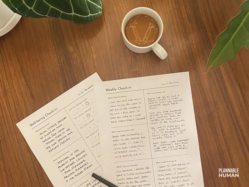 May include: Two printable worksheets on a wooden table with a cup of coffee. The worksheets are titled "Well-being Check-in" and "Weekly Check-in". The worksheets have handwritten text on them.