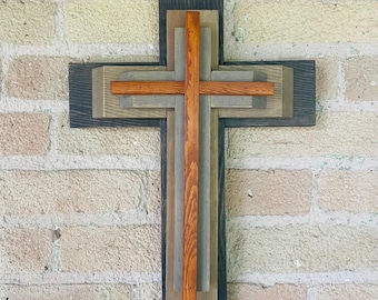 May include: A wooden cross made of multiple layers of wood in shades of brown, gray, and orange. The cross is mounted on a brick wall.