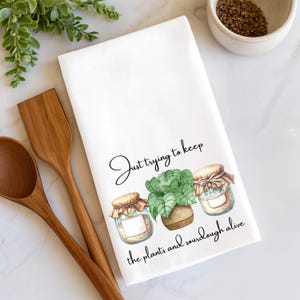 May include: White kitchen towel with the text "Just trying to keep the plants and sourdough alive." The towel features illustrations of a potted plant and two jars with lids. Wooden spoons and a small bowl are in the background.