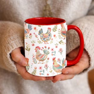 Puede incluir: Una taza de cerámica blanca con interior y asa rojos. La taza está decorada con un patrón repetido de gallinas coloridas, flores y follaje. La taza está sostenida por una persona.