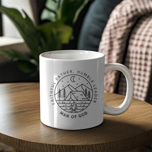 Może przedstawiać: Biały kubek ceramiczny z czarno-białym wzorem graficznym. Wz&oacute;r przedstawia g&oacute;rską scenerię z namiotem, drzewami, ogniskiem i rosnącym księżycem. Tekst wok&oacute;ł wzoru brzmi "Wierny Ojciec, Skromny Przyw&oacute;dca, Człowiek Boga."