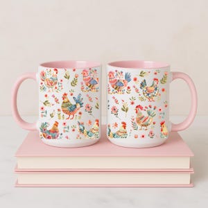 Puede incluir: Dos tazas de cer&aacute;mica blancas con interior y asa rosas. Las tazas presentan un colorido estampado floral y de gallinas. Las tazas est&aacute;n sobre una pila de libros rosas.