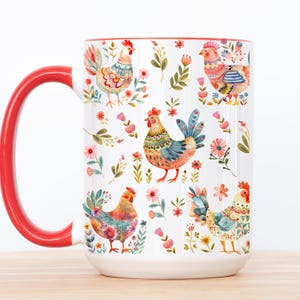 Puede incluir: Taza de cerámica blanca con asa y borde rojos. La taza está decorada con ilustraciones coloridas de gallos y motivos florales. Los diseños de gallos presentan varios colores, incluyendo rojo, azul, amarillo y verde.