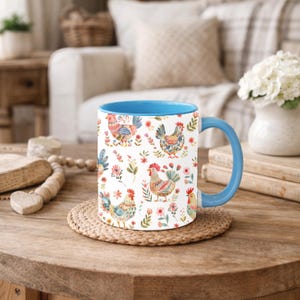 Puede incluir: Una taza de cer&aacute;mica con asa e interior azules. El exterior presenta un patr&oacute;n repetido de gallinas coloridas y elementos florales sobre un fondo blanco. La taza est&aacute; sobre un posavasos redondo tejido sobre una mesa de madera.