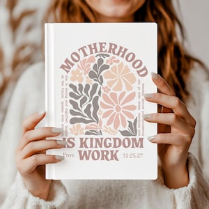 May include: A white journal with the words "MOTHERHOOD" and "IS KINGDOM WORK" in a floral design. The journal includes text from Proverbs 31:25-27. The design features flowers in shades of pink, peach, and gray.