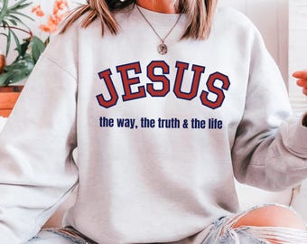 May include: A light gray sweatshirt with the word "JESUS" in red block letters with a navy blue outline. Below the word "JESUS" is the text "the way, the truth & the life" in navy blue.