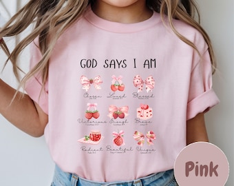 Puede incluir: Camiseta rosa con el texto "GOD SAYS I AM" y varias palabras e ilustraciones inspiradoras. El diseño incluye lazos, fresas y palabras como "Chosen", "Loved" y "Blessed".