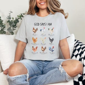 May include: A gray t-shirt with the text "God Says I Am" printed on the front. The text is surrounded by a series of watercolor illustrations of chickens, each with a different word underneath, such as "Chosen", "Loved", "Blessed", "Victorious", "Enough", "Brave", "Radiant", "Beautiful", and "Unified".
