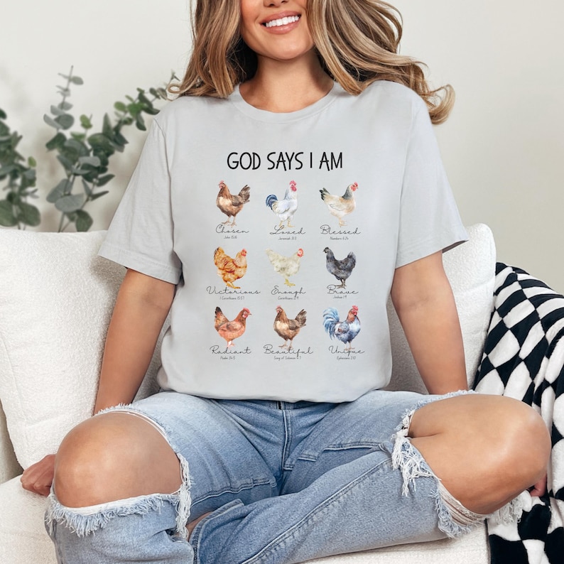 May include: A gray t-shirt with the text "God Says I Am" printed on the front. The text is surrounded by a series of watercolor illustrations of chickens, each with a different word underneath, such as "Chosen", "Loved", "Blessed", "Victorious", "Enough", "Brave", "Radiant", "Beautiful", and "Unified".