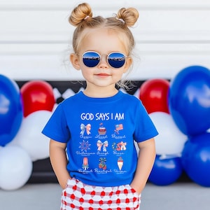 Op de afbeelding: Een kind draagt een blauw t-shirt met de tekst "GOD SAYS I AM" en diverse afbeeldingen. Het kind draagt ook een zonnebril en een rood-wit geruite korte broek. Rode, witte en blauwe ballonnen zijn op de achtergrond te zien.