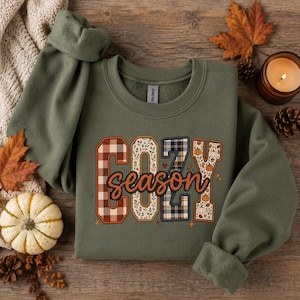 May include: An olive green crewneck sweatshirt featuring the text COZY season printed on the front. The letters are filled with various patterns including red and white plaid, floral motifs, and navy blue plaid, with orange pumpkin graphics.