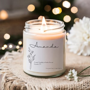 Puede incluir: Una vela blanca en un tarro de vidrio con una etiqueta que dice "Amanda" y un diseño floral. La etiqueta también incluye la cita "Una dulce amistad renueva el alma" y la referencia bíblica "Proverbios 27:9".