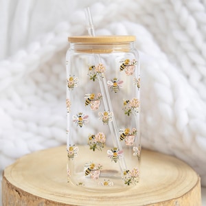 Könnte beinhalten: Ein durchsichtiger Glasbecher mit Bambusdeckel und Strohhalm. Der Becher ist mit einem sich wiederholenden Muster aus Bienen und rosa Blumen verziert. Der Becher steht auf einer Holzoberfläche.